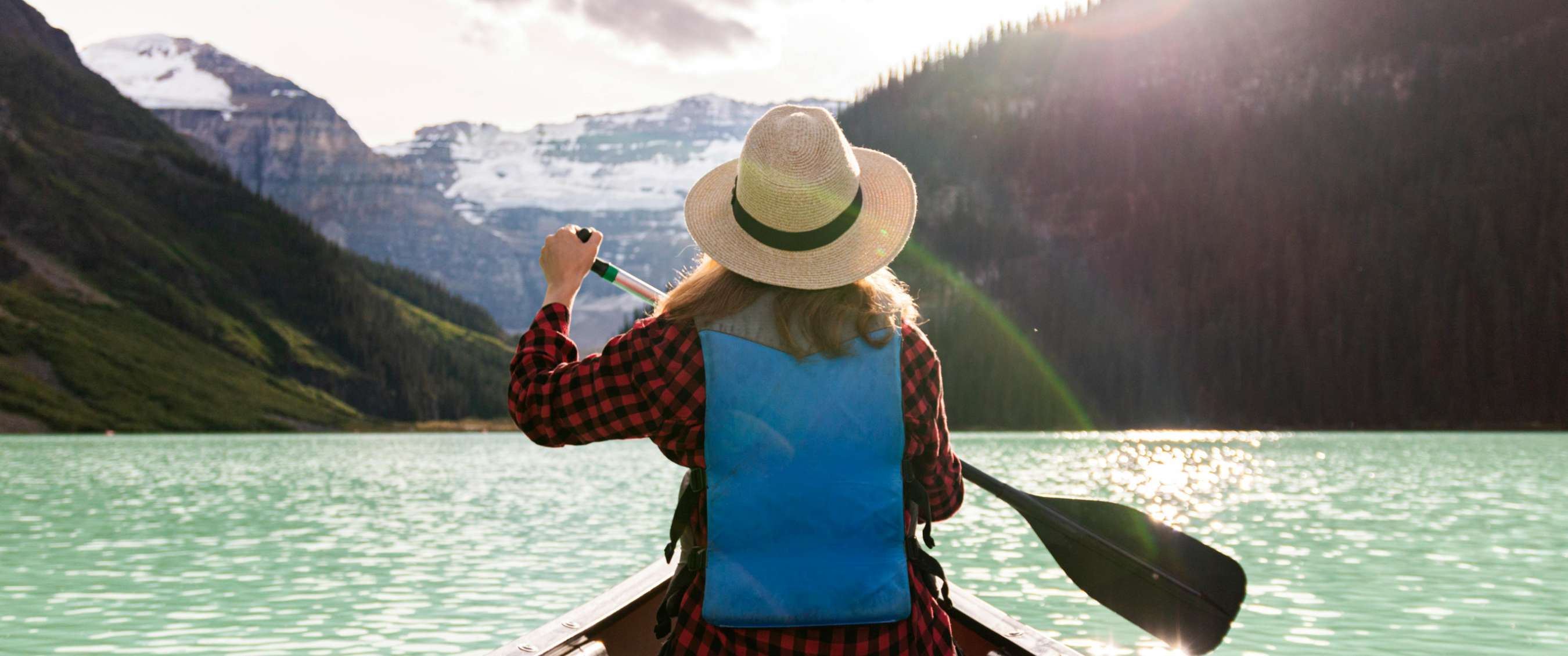 Vrouw met hoed en ruitjeshemd peddelt op turquoise bergmeer richting besneeuwde bergen.
