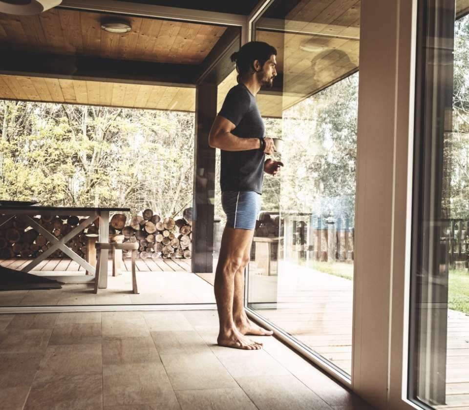Jonge man staat gekleed in thermisch ondergoed door het grote raam van een woning naar de bosrijke omgeving te kijken.