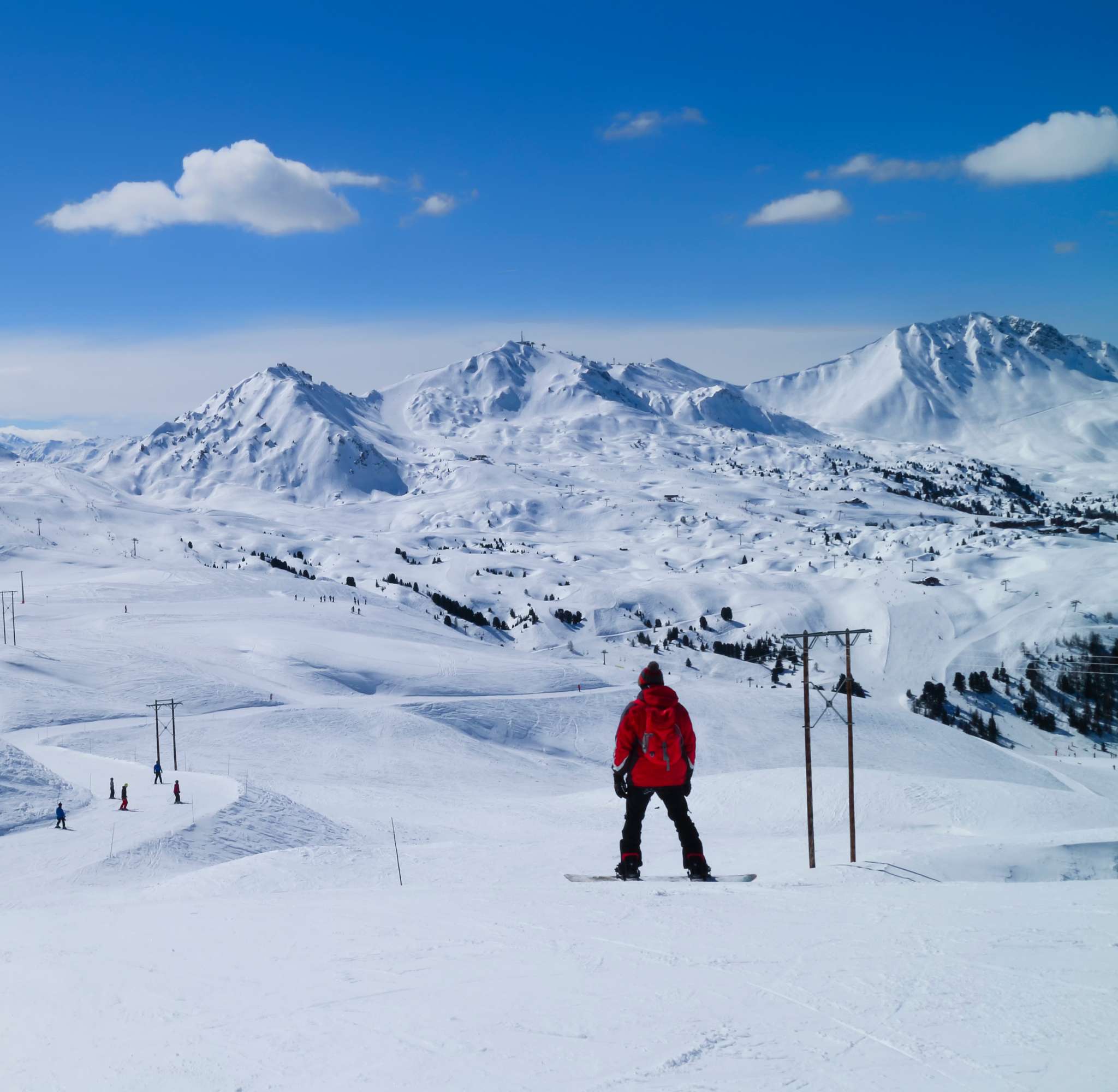 Een snowboarder in een rode jas staat op een besneeuwde helling met uitzicht op een uitgestrekt berglandschap in La Plagne.